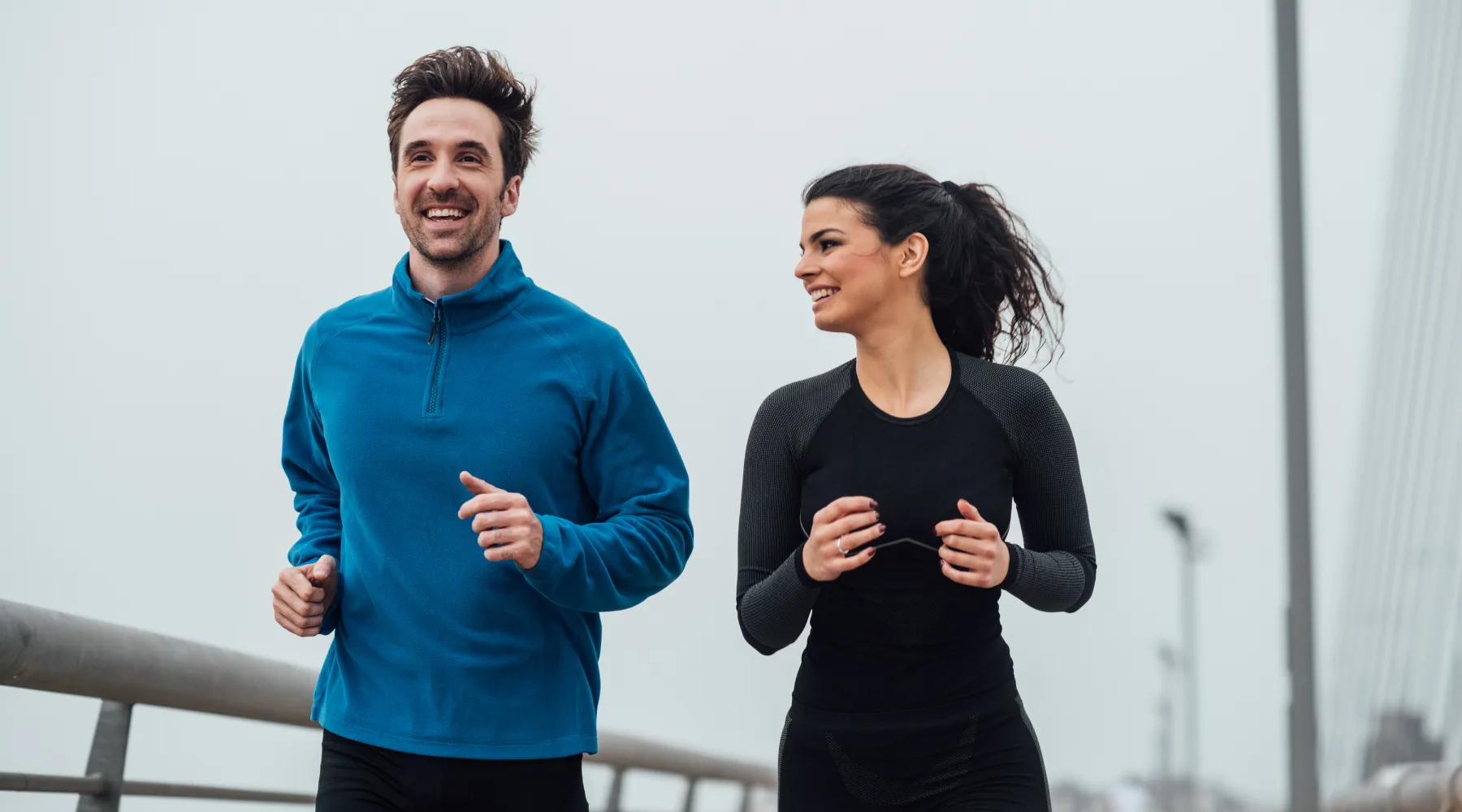 A couple jogging together outdoors on a bridge, smiling as they run side by side
