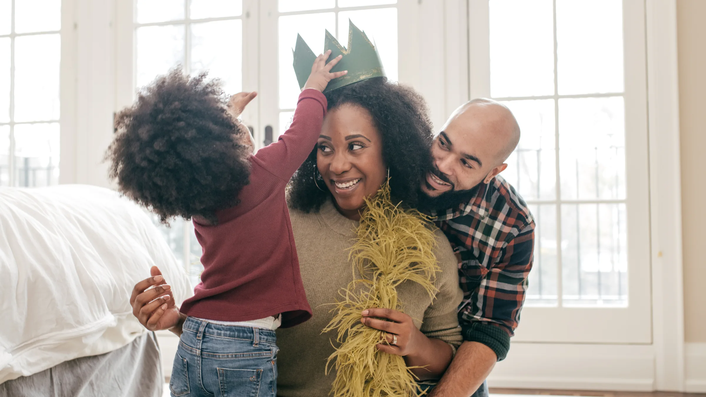A family sharing a joyful moment at home — a child playfully placing a paper crown on their mother's head while the father laughs alongside them