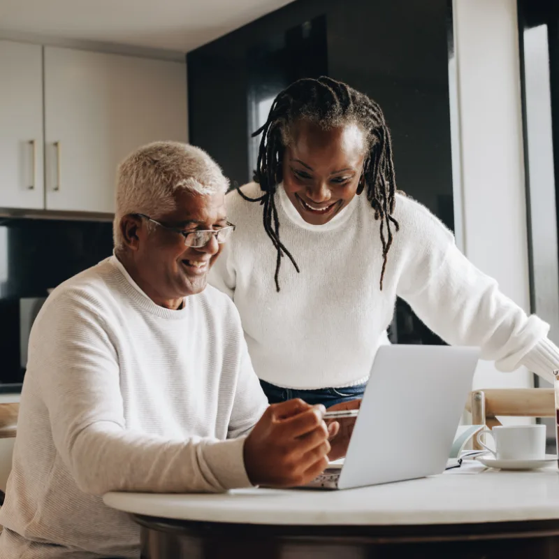 A couple sitting at their kitchen table together, looking at a laptop and reviewing their finances with confidence