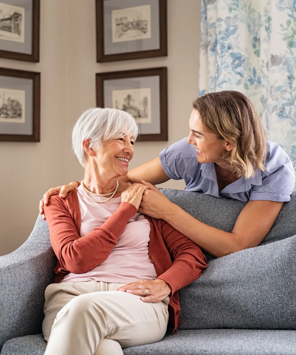 A compassionate caregiver sitting beside an older woman on a couch, both smiling gently — representing the living benefits that let policyholders access their coverage during serious illness or injury