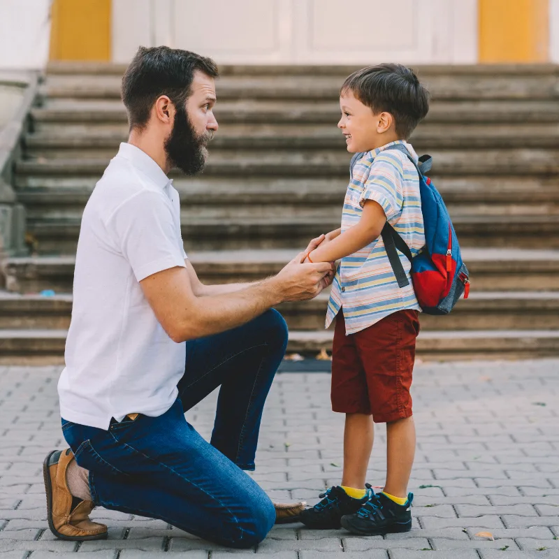 A father kneeling to help his young son put on a school backpack, representing the affordable family protection that term life insurance provides during key earning years