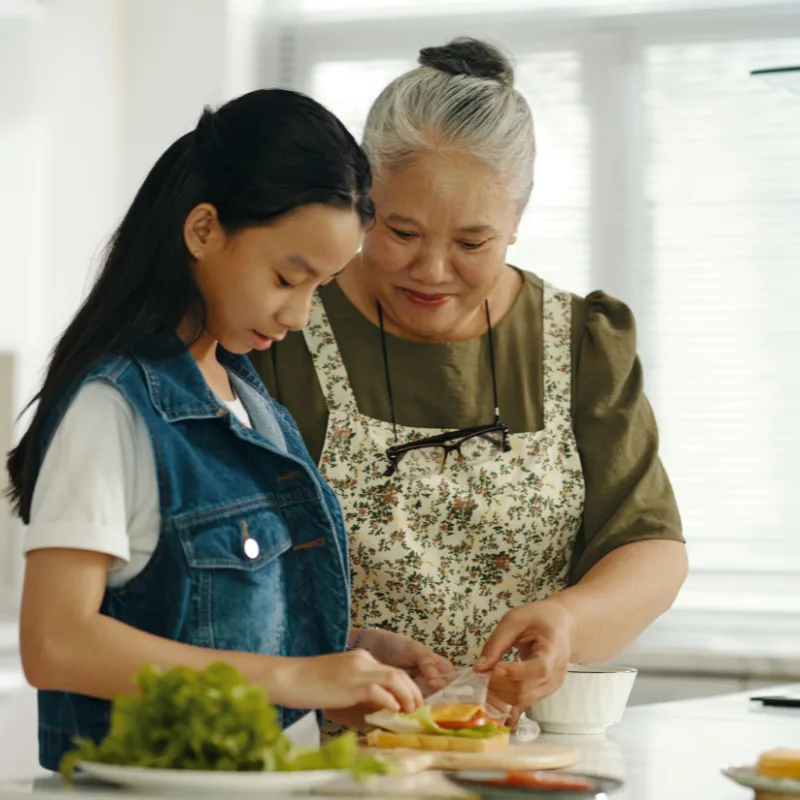 A grandmother and granddaughter smiling warmly together in the kitchen while making sandwiches, representing the generational legacy and permanence of whole life insurance