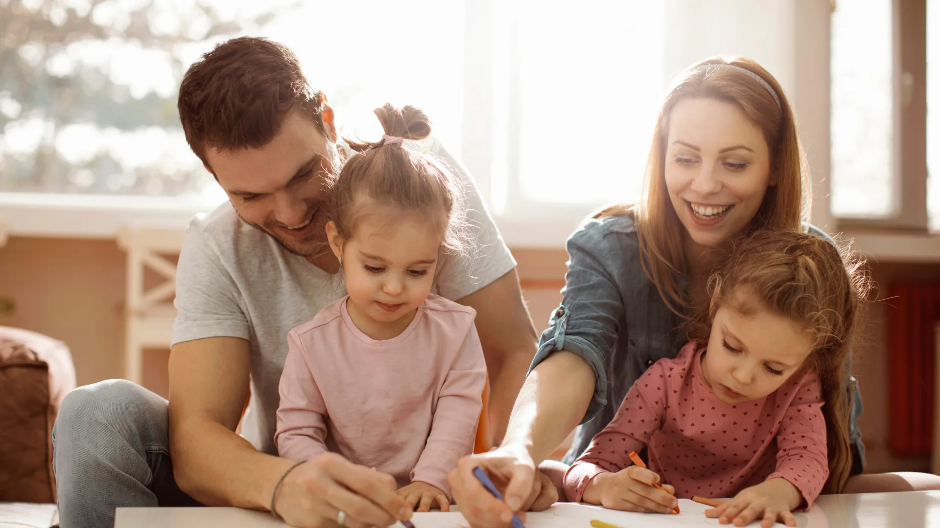 A family of four — two parents and two young daughters — coloring together at a sunlit table, representing the peace of mind that comes with supplemental health coverage