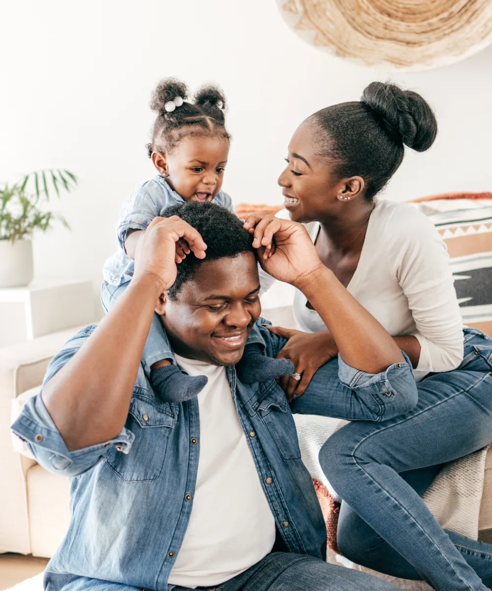 A dad with a baby on his shoulders while mom plays with them — a joyful family moment representing the protection whole life provides